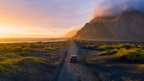 A single car drives along a lonely gravel road in golden sunlight, surrounded by mountains and open countryside A single car drives along a lonely gravel road in golden sunlight, surrounded by mountains and open countryside